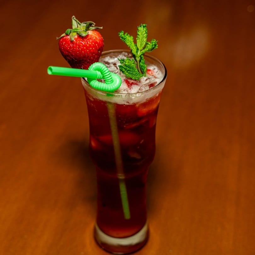 A tall glass of iced red drink with a strawberry, mint, and green straw.