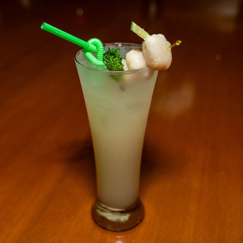 A tall glass of lychee martini cocktail with a green straw, mint, and lychee garnish.