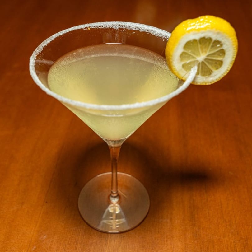 A lemon drop martini with a sugared rim and a lemon slice garnish on a wooden table.