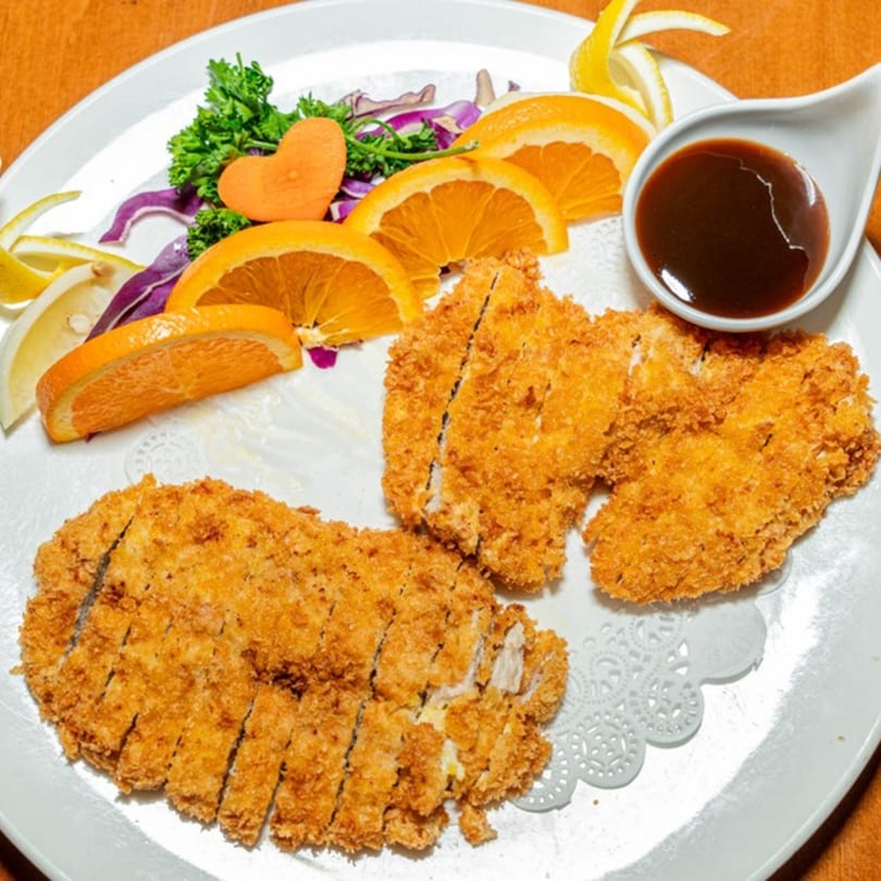 Crispy fried chicken cutlets with orange slices, parsley, and a side of dark sauce on a white plate.
