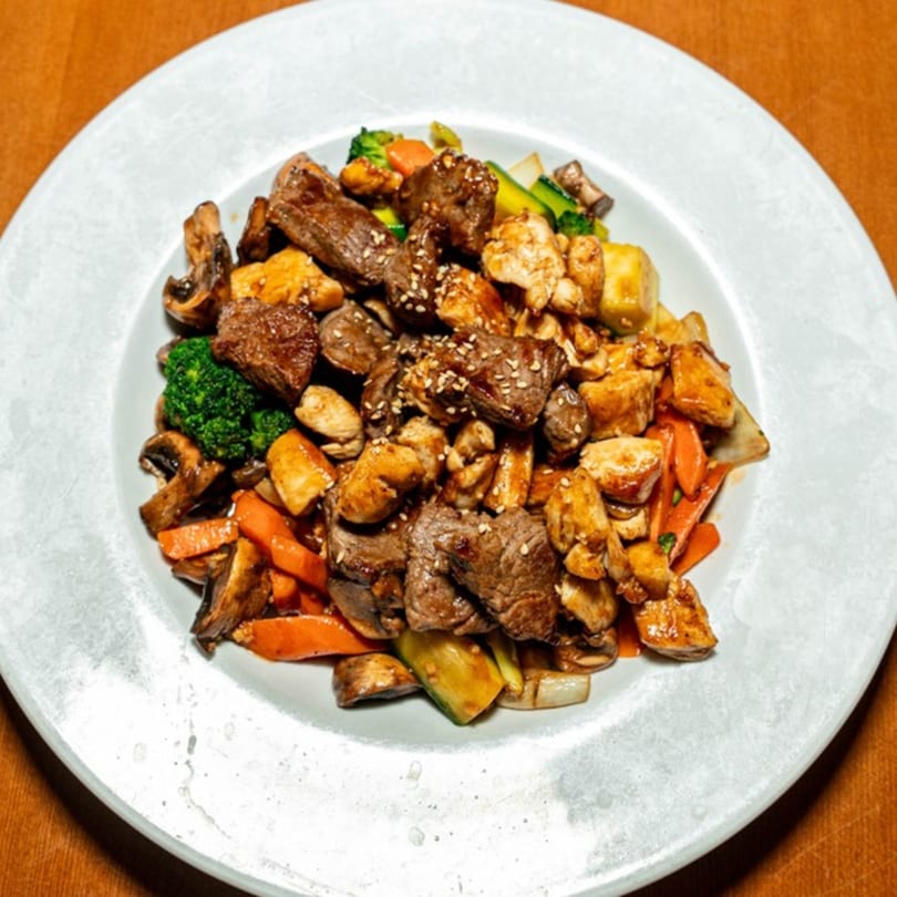 A white plate filled with stir-fried beef, chicken, mushrooms, broccoli, and carrots, topped with sesame seeds.